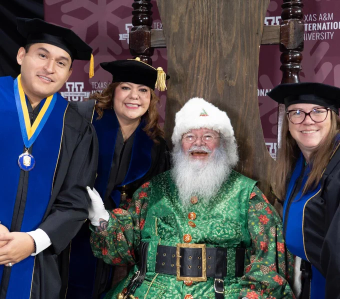 Four individuals pose for a photo at a graduation event, including three people wearing academic regalia and Santa Claus. They stand in front of a Texas A&M International University backdrop, smiling toward the camera.