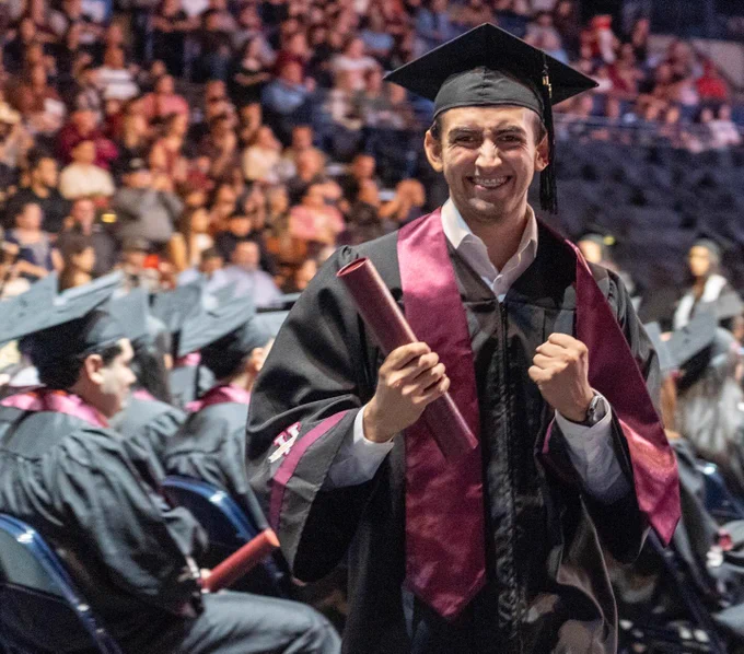 A graduate in a black cap and gown smiles and raises both fists in celebration while holding a diploma on the commencement floor. Rows of seated graduates and audience members fill the arena behind them.