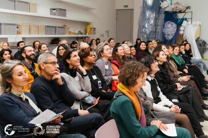 
People seated in rows at the Multaka 10th anniversary. In the background, there are shelves with boxes and a large sculpture.
