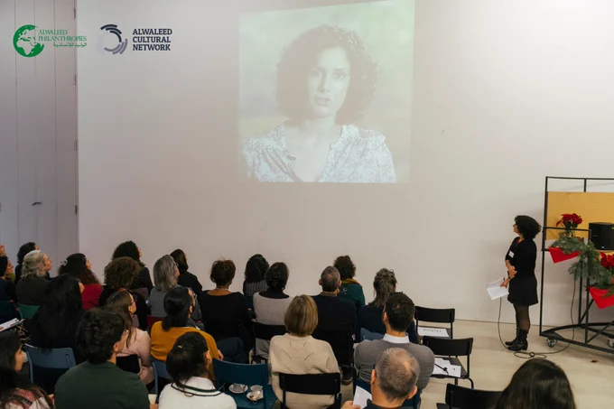 An audience seated in rows at the Multaka Anniversary event watching a video. The video features a person who is speaking.
