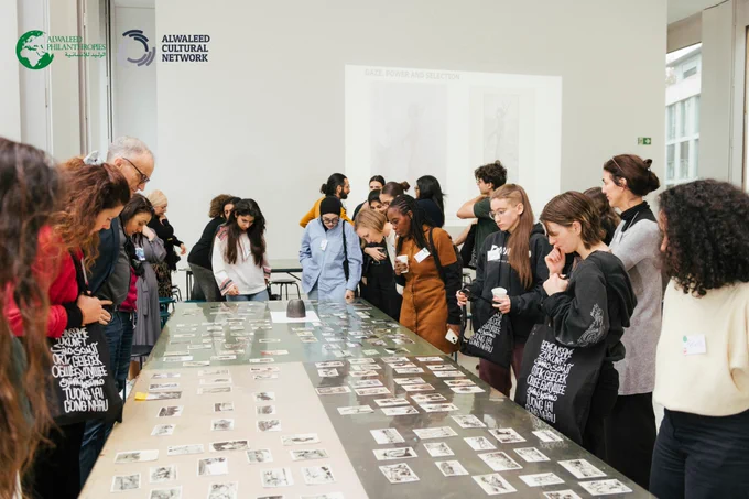 Workshop as part of the Multaka Anniversary event. There are photographs laid out on a glass table as part of an exhibition. People are standing around the photographs, looking at them, and discussing them.