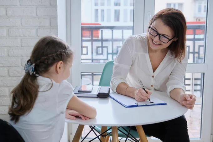 a smiling woman sits at a child's table taking notes while talking girl