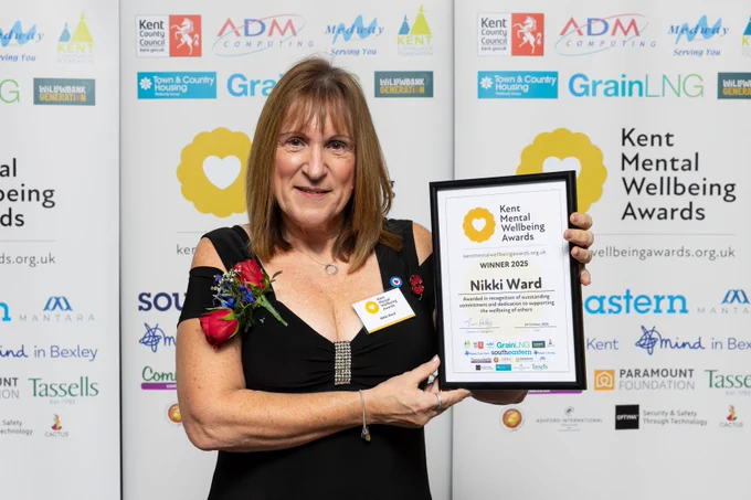 Nikki is stood in front of a Kent Mental Wellbeing Awards banner. She is smiling as she holds up her winner's certificate