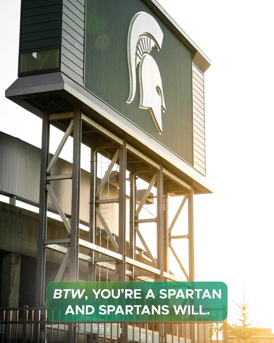 A view looking up at the Spartan head logo on top of a building with a metal frame. In the foreground, a green text box reads, "BTW, YOU'RE A SPARTAN AND SPARTANS WILL."