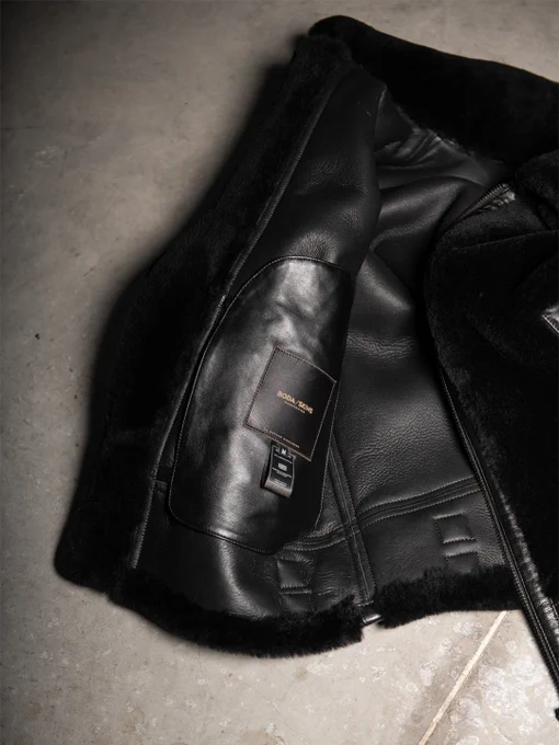 Interior detail of a black shearling and leather jacket showing textured leather, soft shearling trim, and a stitched brand label.