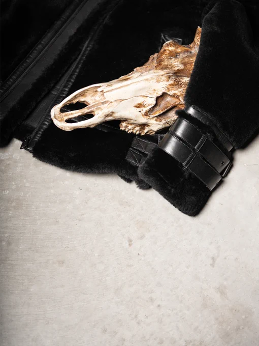 Detail shot highlighting the plush black shearling cuff and leather strap of a jacket, contrasted with an animal skull on a neutral concrete surface.