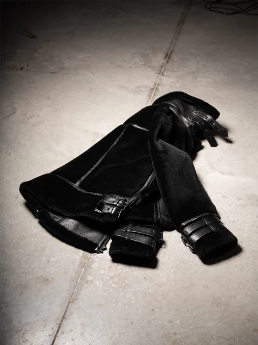 Flat-lay of a black shearling jacket showcasing plush texture, leather trim, and buckle accents against a neutral concrete surface.