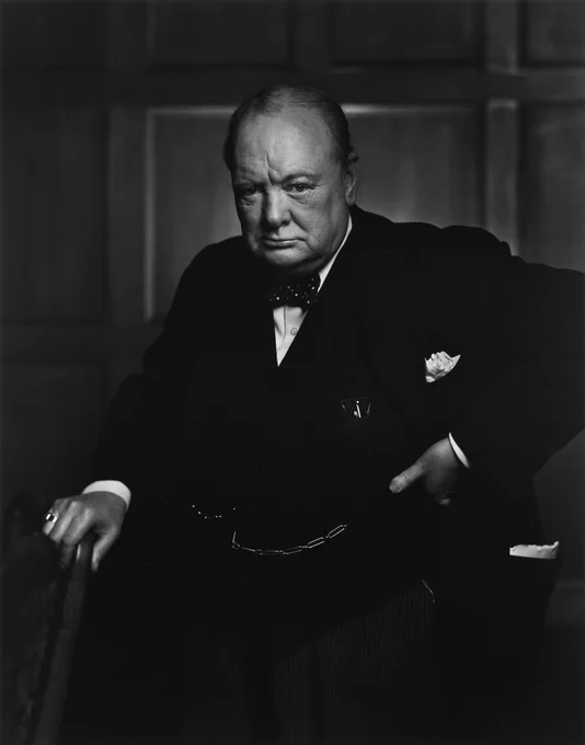 Image description: Black and white archive photograph of Sir Winston Churchill looking at the camera. He is wearing a suit with white-spotted bowtie, a chain across his waistcoat and holding a cane with his right hand.
