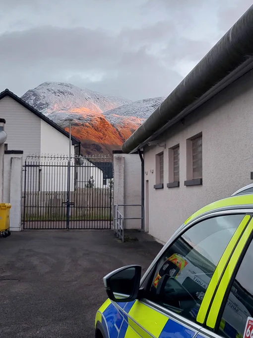 A Police Scotland vehicle parked beside buildings at a police station, with the snow dusted Ben Nevis and hills visible in the background.