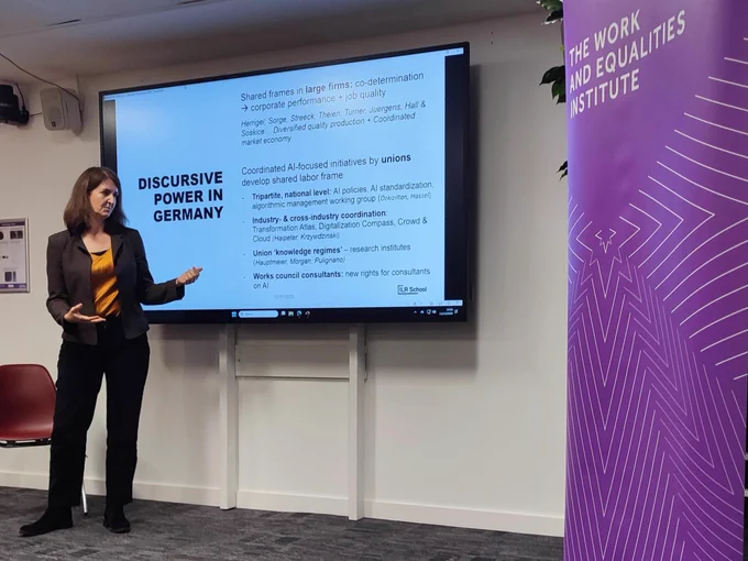 A woman presenting at a screen with a purple banner to the right of the image.