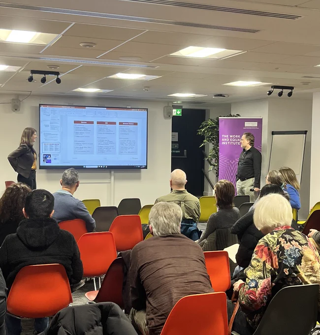 A woman presenting to a group of people in a seminar room. A man stands on the right in front of a purple banner.