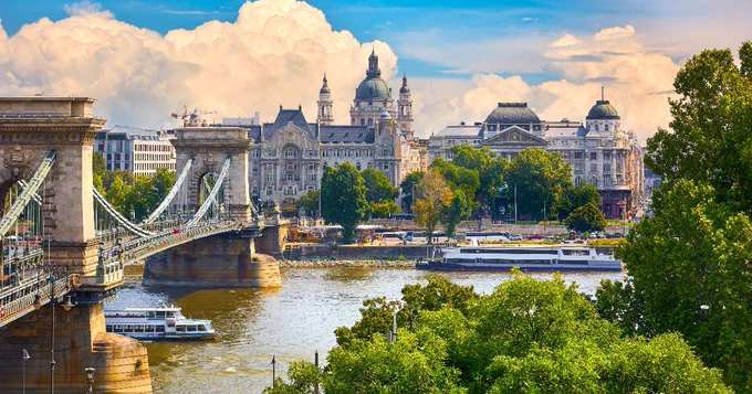 View of Budapest featuring the Chain Bridge over the Danube River, with the Gresham Palace in the background under a cloudy sky.