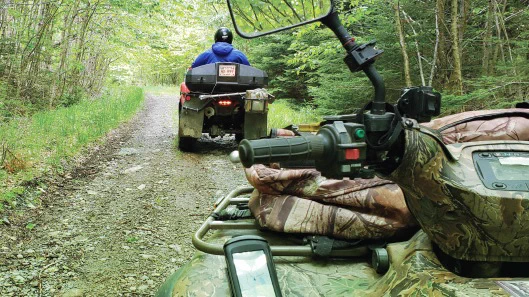 Two ATVs drive down a dirt and gravel trail in the woods. 