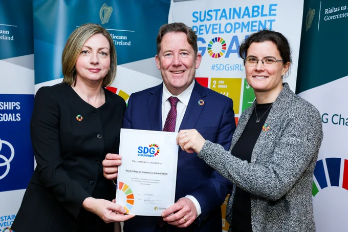 Three people stand together at an event backdrop featuring Sustainable Development Goals branding. They are holding a certificate that reads “SDG Champion,” awarded to the Royal College of Surgeons in Ireland (RCSI). All three are wearing SDG lapel pins, and the backdrop includes Irish government and SDG Ireland logos.