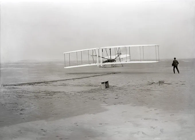 The first powered, controlled, sustained airplane flight in history. Orville Wright, age 32, is at the controls of the machine, lying prone on the lower wing with hips in the cradle which operated the wing-warping mechanism. His brother, Wilbur Wright, age 36, ran alongside to help balance the machine, having just released his hold on the forward upright of the right wing. Image source: Library of Congress