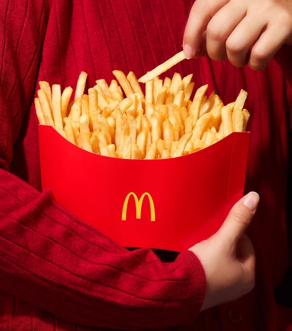 The image shows a close-up of a person holding a large red McDonald’s french fry container filled to the brim with golden, crispy fries. The iconic yellow McDonald’s “M” logo is prominently displayed on the front of the container. The person is wearing a textured, ribbed red sweater, which matches the color of the fry box, creating a visually cohesive look. One hand is cradling the container, while the other is picking up a single fry. The background is out of focus, emphasizing the fries and the hands as the main subjects. The lighting is bright and even, highlighting the warm tones of the fries and the vibrant red of the packaging