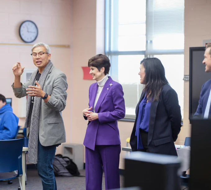 A professional group engages in a discussion inside a classroom setting, with one individual speaking animatedly. The atmosphere appears collaborative and focused.