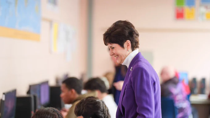 A person in a purple blazer smiling in a computer classroom filled with students.