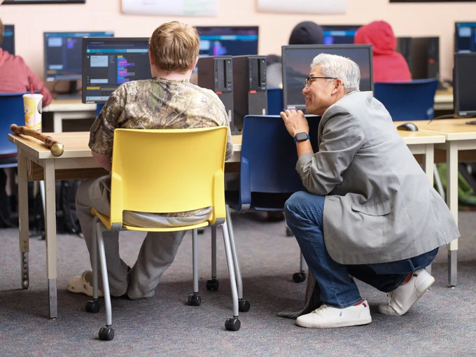 Two individuals are seated at computers in a classroom setting, one in a camouflage shirt and the other dressed in a gray blazer, closely observing the screen.