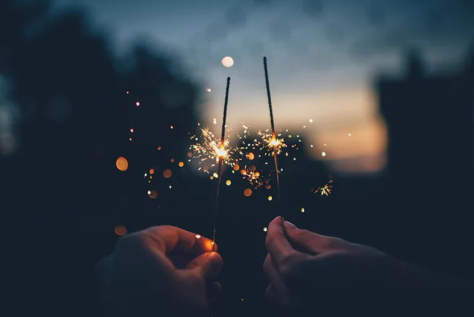 The hands of two people hold sparklers side by side. In the distance, we see the twilight horizon.
