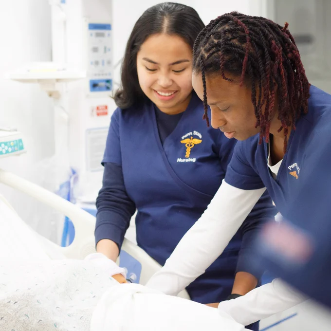 two students wearing penn state nursing uniforms work on a patient simulator in a hospital setting