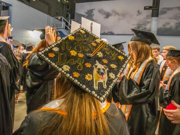 Graduation cap decorated with the phrase "Did it for my dog" and an image of a bulldog, worn by a student in a crowd of graduates.