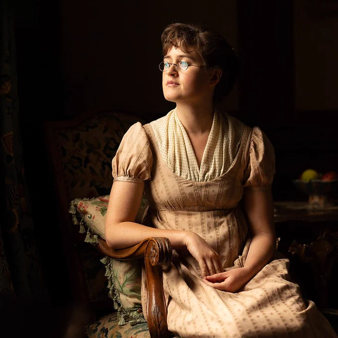 A young woman in a pale dress sits in a chair, looking out a window