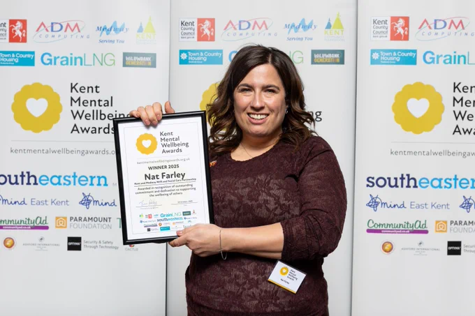 Nat Farley is stood, smiling in front of a Kent Mental Wellbeing Awards banner holding up her winner's certificate