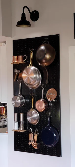 Black metal Wall Control pegboard mounted on a kitchen wall, holding neatly organized pots, pans, copper cookware, spice grinders, and utensils under a wall-mounted light.