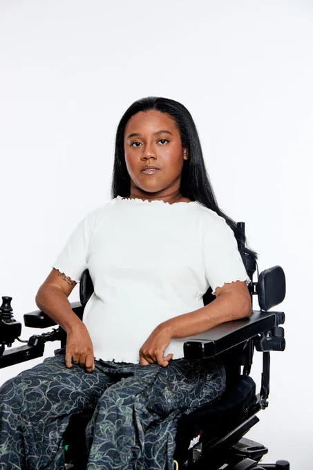 A young woman with long straight black hair and medium-dark skin sits in a black motorized wheelchair, wearing a white short-sleeve top with frayed edges and patterned green pants against a white background. She has a tattoo on her right forearm.
