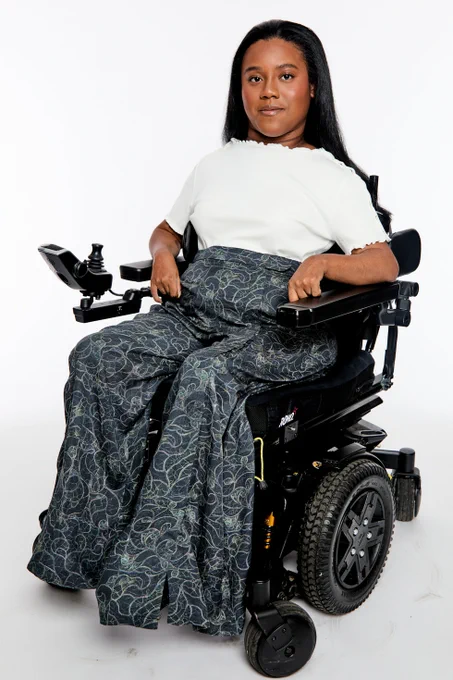 A woman with long black hair, wearing a white short-sleeve top and a long dark pants with a light green swirl pattern, sits in a black powered wheelchair with joystick control, positioned against a white background.