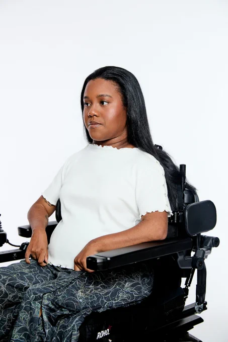 A woman with long black hair and medium-dark skin, wearing a white short-sleeve shirt with scalloped edges and a patterned green pants, sits in a black powered wheelchair with the brand “ROHO” visible on the cushion, looking to the side against a white background.