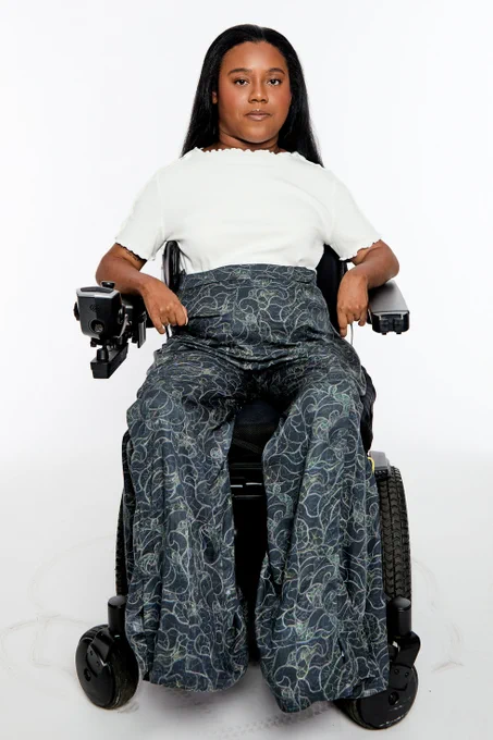 A woman with long black hair, wearing a white short-sleeve shirt and dark patterned pants, sits in a black manual wheelchair against a white background.