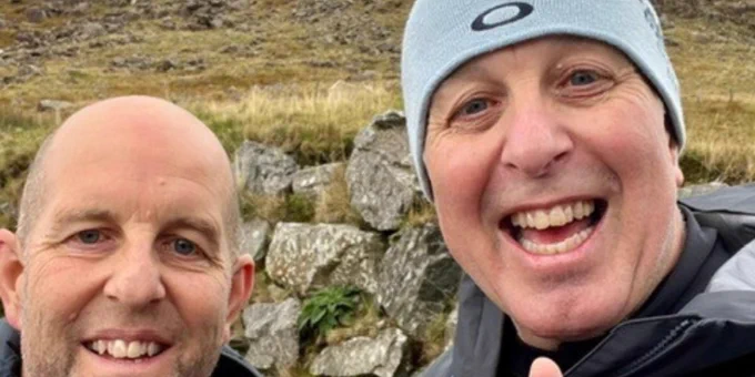 Barrie and his brother David giving thumbs up with rocky hillside and cloudy sky in the background.