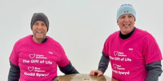 Barrie and his brother David wearing bright pink 'The Gift of Life' shirts stand beside a stone trig point on a foggy mountaintop.