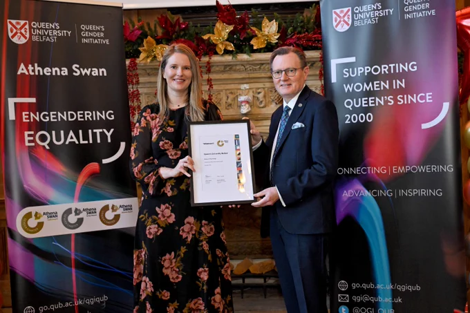 Two individuals holding a framed certificate, standing between banners promoting Queen's University Belfast's Gender Initiative and Athena Swan equality program.