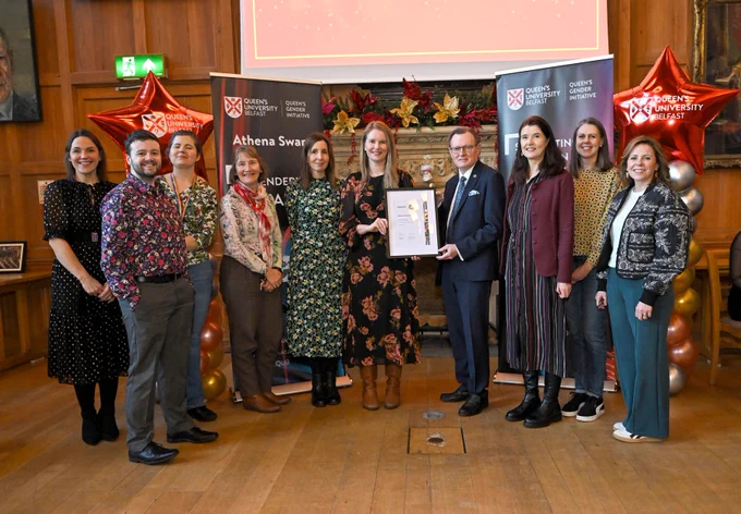 Group of nine people standing indoors at Queen's University Belfast event, one holding a framed certificate with celebratory balloons in the background.