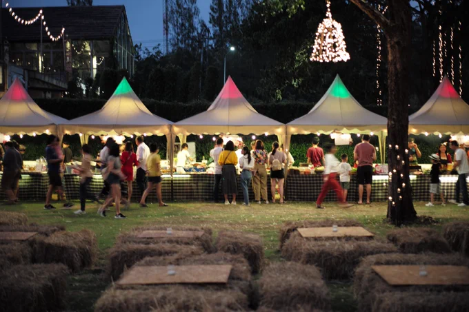 People walking through an outdoor food market at night, with illuminated tents, string lights, and hay bales arranged as seating.