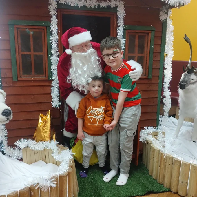 Two children posing with Santa Claus in a festive winter cabin decorated with white garlands and reindeer figures.