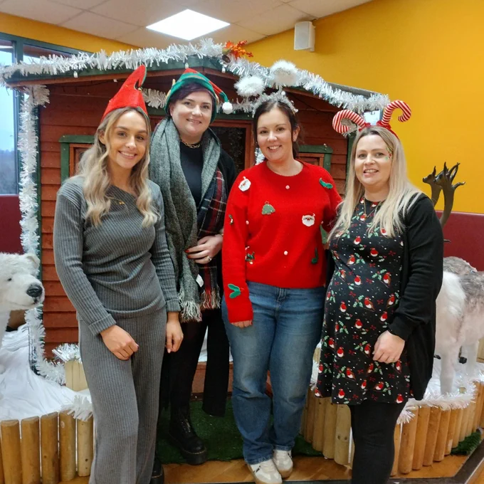 Four women dressed in festive holiday attire stand in front of a wooden cabin decorated with white garlands and surrounded by reindeer figures.