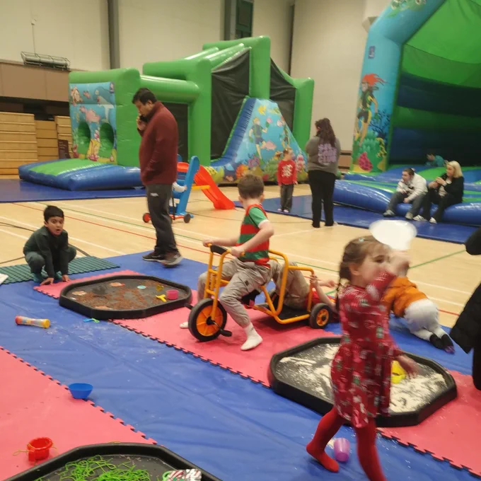 Children playing on colourful mats with sensory trays and riding toys inside a gymnasium with inflatable bouncy castles in the background.