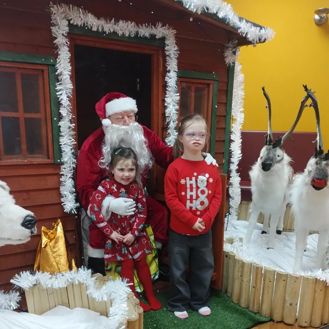 Children posing with Santa Claus outside a festive wooden cabin decorated with white garlands and surrounded by reindeer figures.