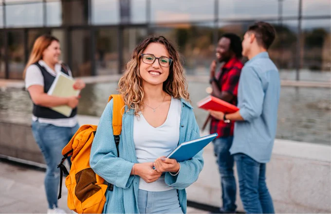 A group of four people standing outdoors near a modern building with large glass windows and a water feature. The person in the foreground is wearing a light blue jacket, white top, and jeans, holding a blue notebook and carrying a bright yellow backpack. In the background, three others are holding books and appear to be engaged in conversation. The setting suggests a campus or academic environment.