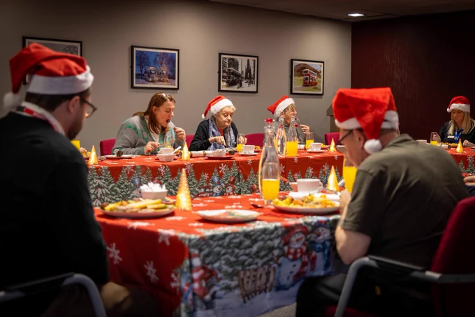 Group of people wearing Santa hats enjoying a festive meal around a table with Christmas-themed decorations and food.