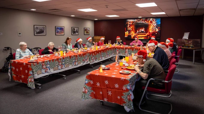 Visitors wearing Santa hats gathered around a U-shaped table decorated with red, festive tablecloths, enjoying a Christmas meal in a cosy room with a fireplace on the tv screen.