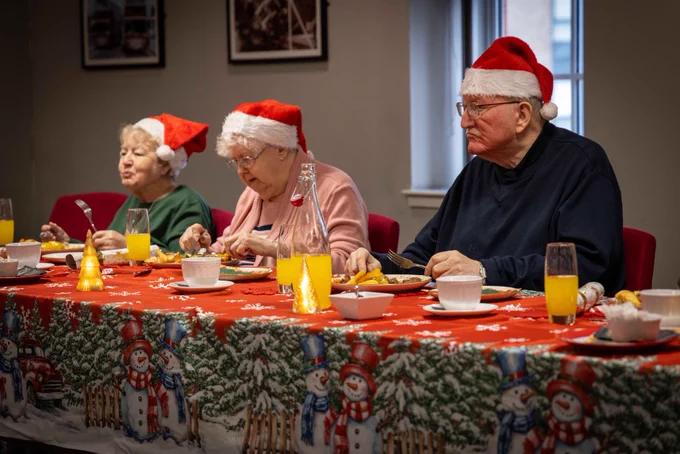 Three elderly people wearing Santa hats enjoy a festive meal at a table decorated with a Christmas-themed tablecloth and orange juice glasses.