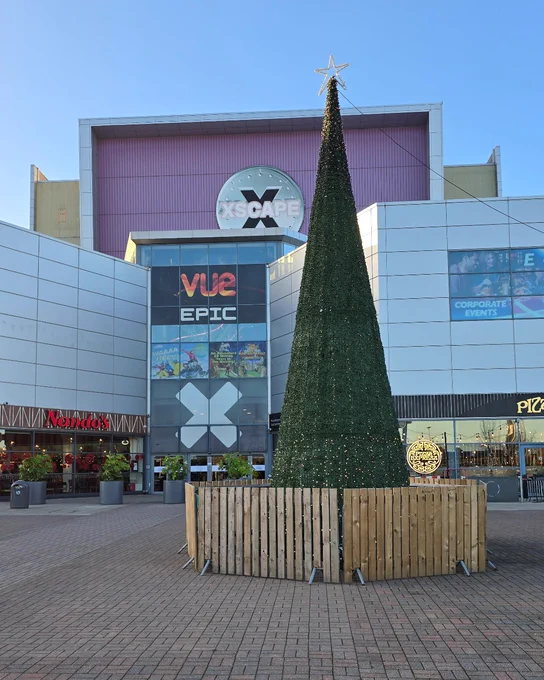 Large Christmas tree decorated with lights and topped with a star, displayed in front of the Xscape building with visible signage for Vue and other businesses.