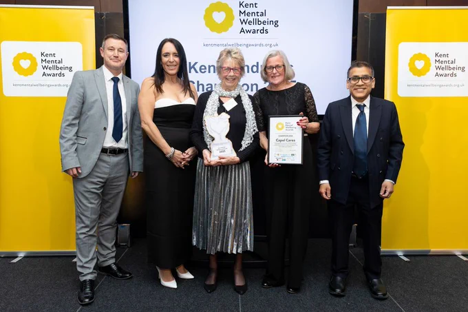 Staff from Capel Cares are holding the winner's certificate and the champions trophy. They're stood in front of a Kent Mental Wellbeing Awards banner with Lee Woolcott-Ellis from Southeastern, Natalie Wyborn from Guildcrest Estates Ltd, and our VIP speaker Dr Kush Kanodia