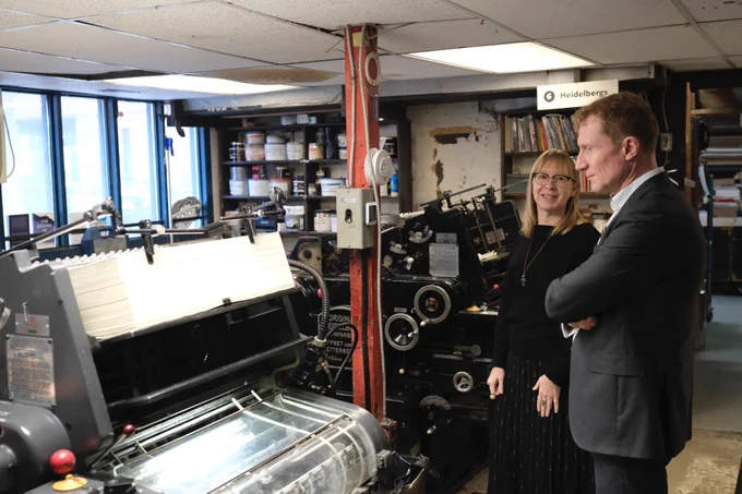 Minister of Canadian Identity and Culture, Marc Miller, views a Heidelberg KORD offset printing press in the Coach House Books office in Toronto, ON, accompanied by Coach House editorial director, Alana Wilcox. Other historical printing machines are visible in the background.