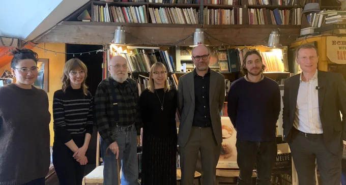 Five Coach House Books staff stand with Association of Canadian Publishers executive director, Jack Illingworth, and Minister of Canadian Identity and Culture, Marc Miller, in the Coach House Books office in Toronto, ON. Books are visible on shelves up to the ceiling and in the background. Everyone is smiling.